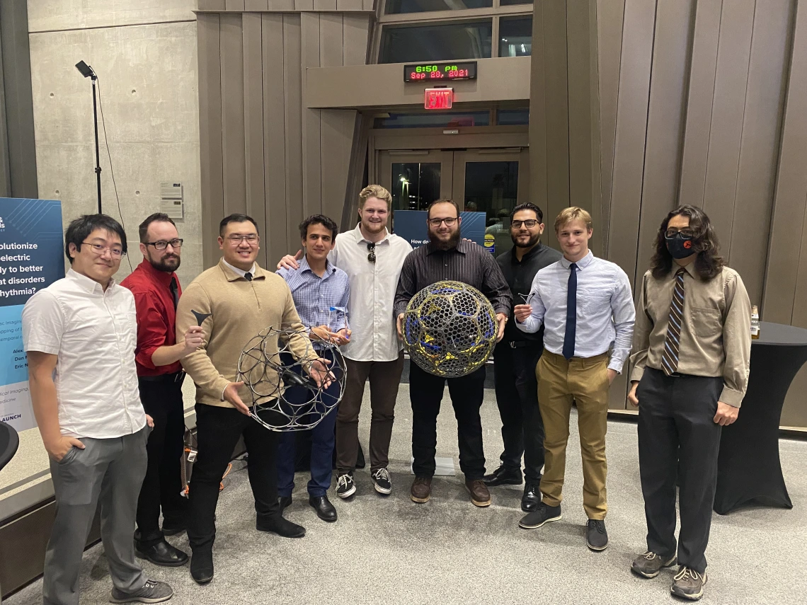 Nine men stand in a row. Two are holding large, spherical mesh robots.