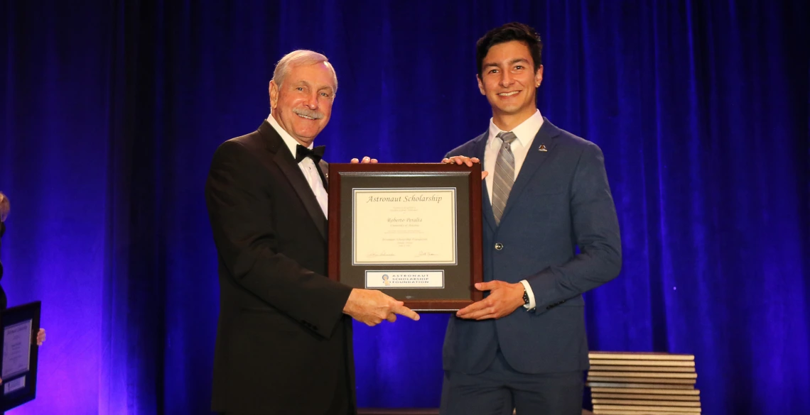 Roberto Peralta stands on a stage accepting a framed certificate from an older man. The certificate says "Astronaut Scholarship." The men are in formal wear and there is a blue curtain behind them.