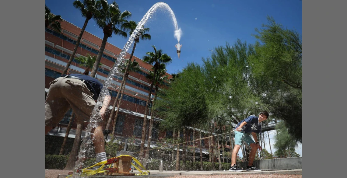 A student pulls on a rope to launch a bottle rock, which is making a big arc through the sky and back toward the ground.