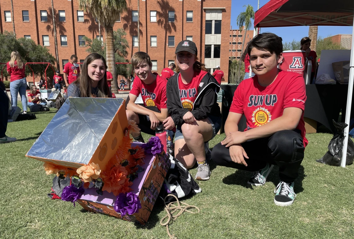 Four students in matching red t-shirts pose on the lawn next to their solar oven -- a cardboard box with a funnel shape on top, decorated with orange and purple tissue paper.