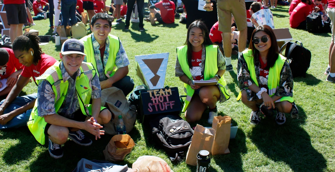 Two young men and two young women wearing Hawaiian shirts and bright yellow vests kneel for a photo next to their solar oven, a cardboard and aluminum foil device labeled with their group name: "Team Hot Stuff."