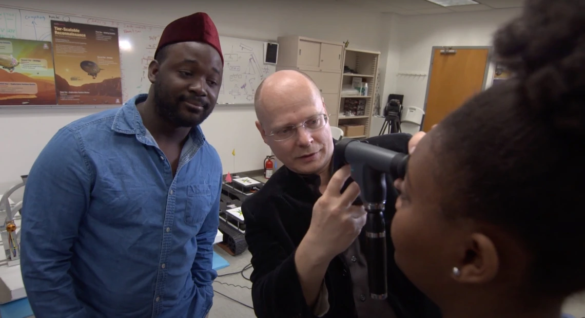 Wolfgang Fink uses an optical device to examine a young woman's eyes, while another young man looks on in the background.