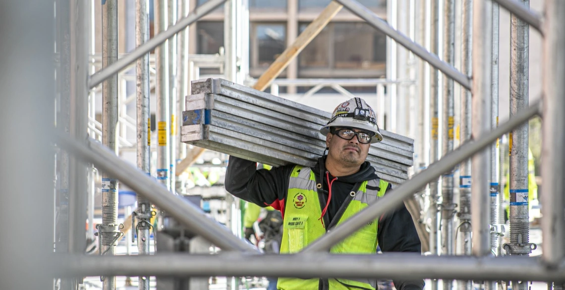 A man wearing a construction vest carries four metal beams on a construction site.