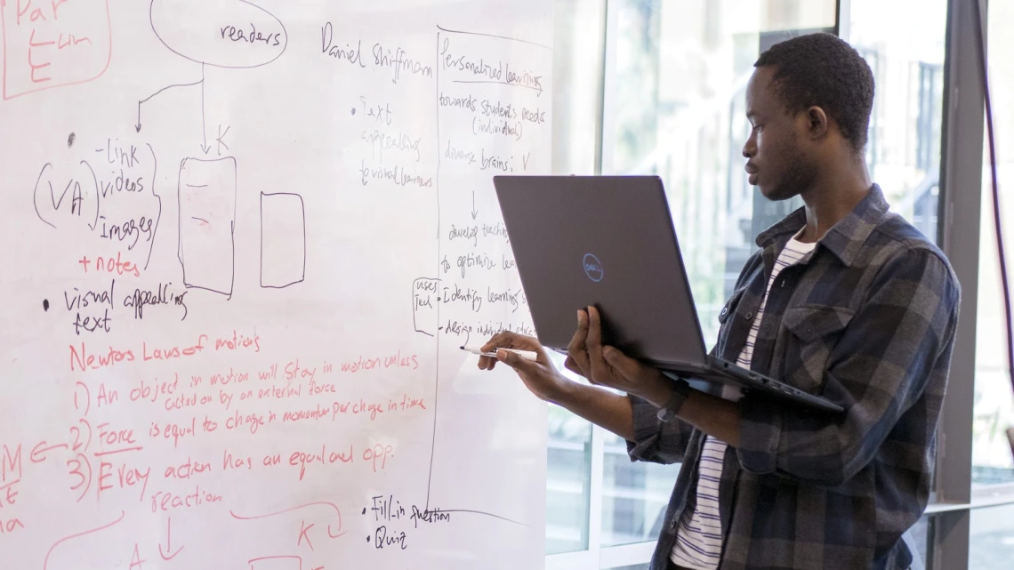 A student holds a laptop and writes on a dry erase board.