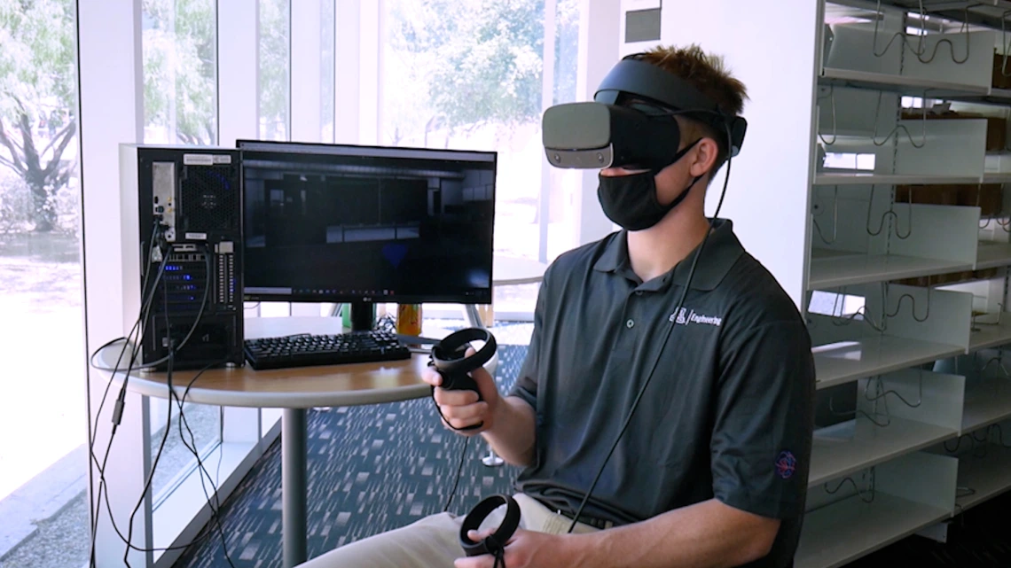 A student sitting next to a computer monitor and wearing a VR headset.