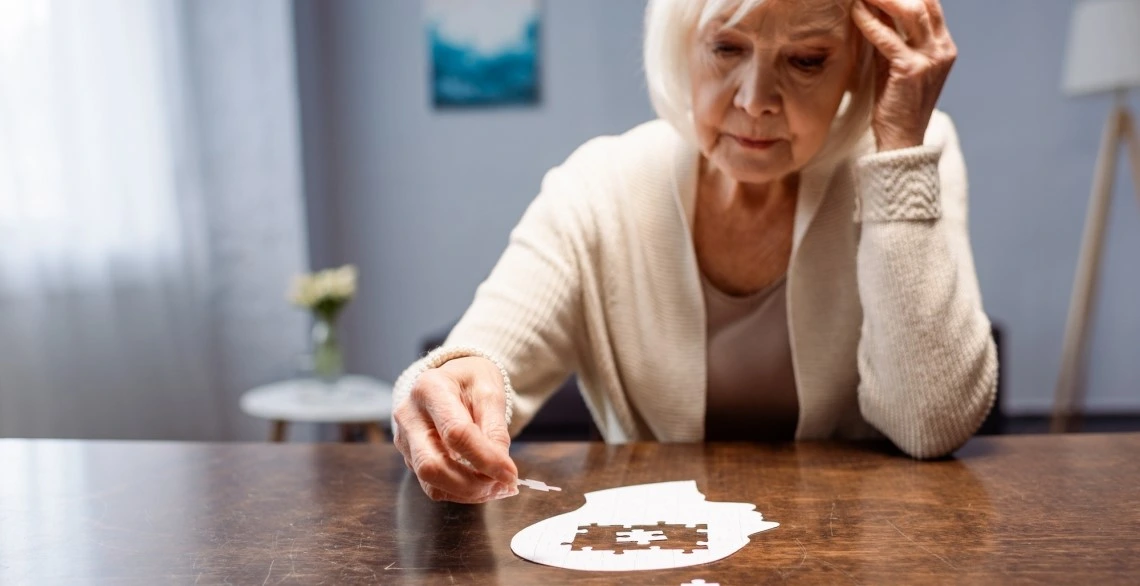 An older woman sitting at a table completing a white puzzle shaped like a human head.