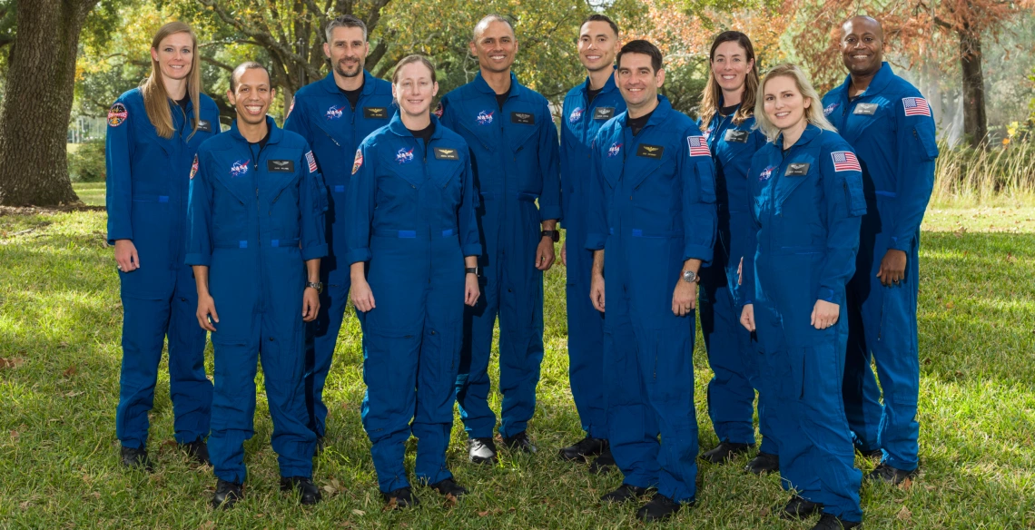 A group of ten astronauts stand in matching blue jumpsuits in a green outdoor area.