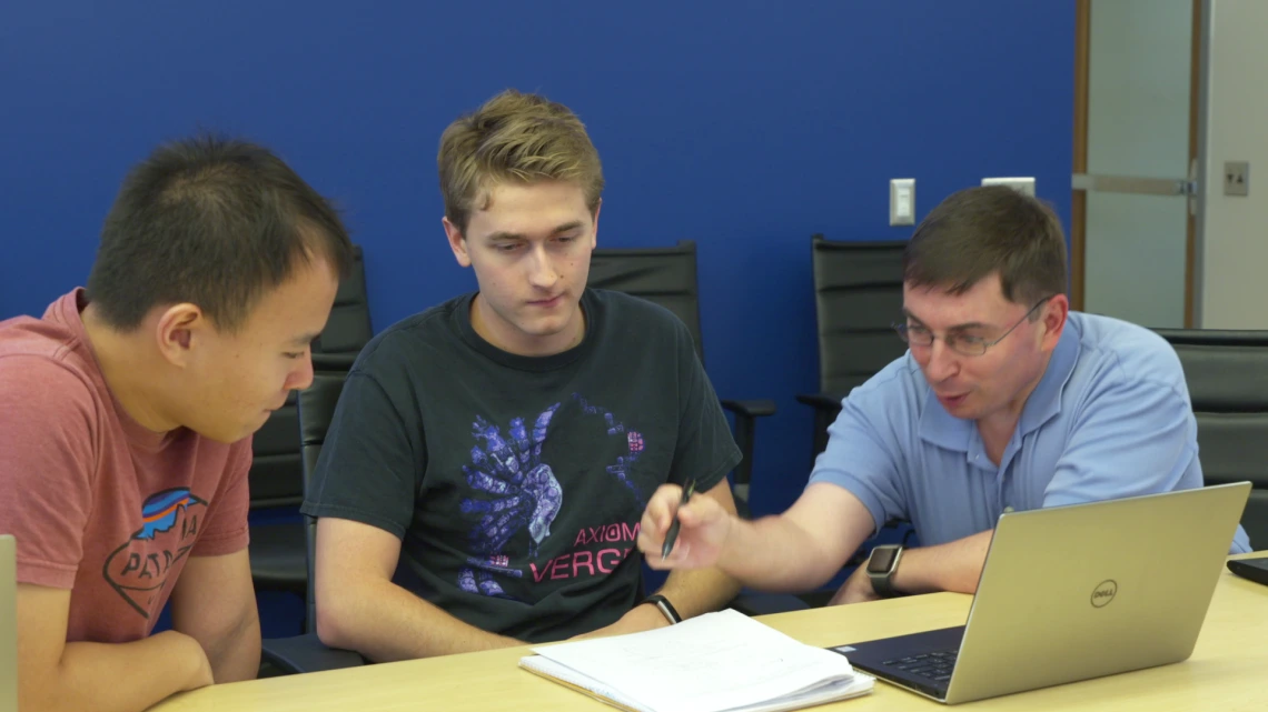 Three men lean over a laptop and set of papers on a table.