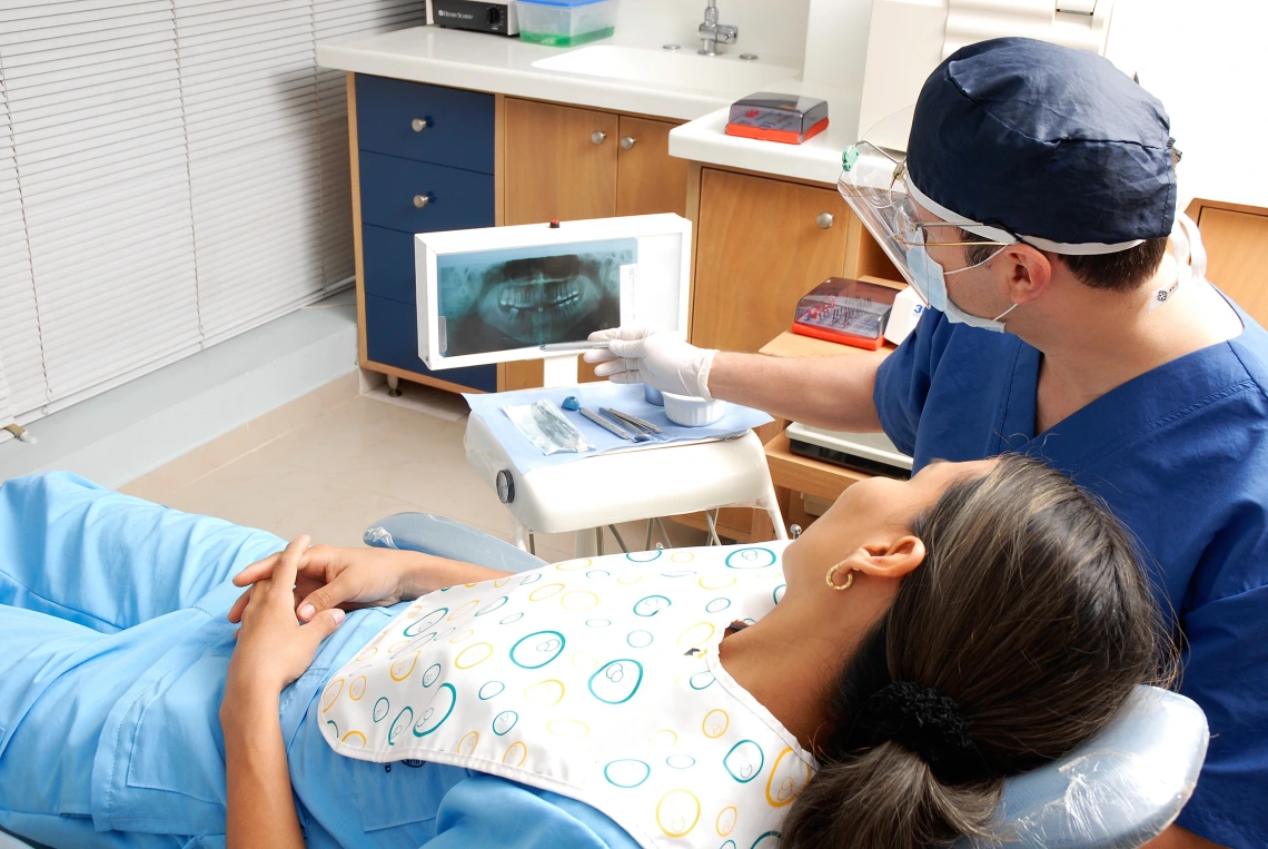 A dental patient lies in a chair while a dentist gestures toward X-rays of the patient's teeth.