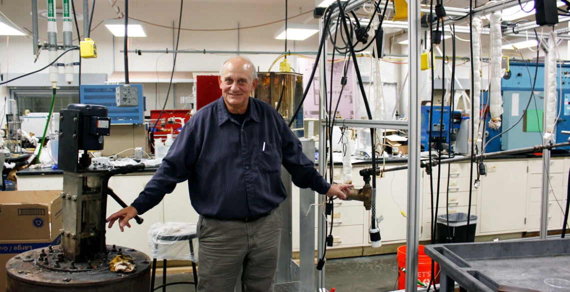 Don Gervasio, wearing a long-sleeved blue shirt, stands between a pump and pipe in a laboratory with tubes hanging in the background