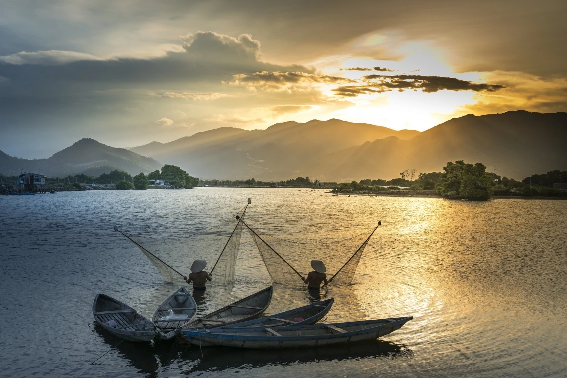 two fishers standing in water and holding up nets. There are four canoes behind them. In the distance is a sunset over mountains.