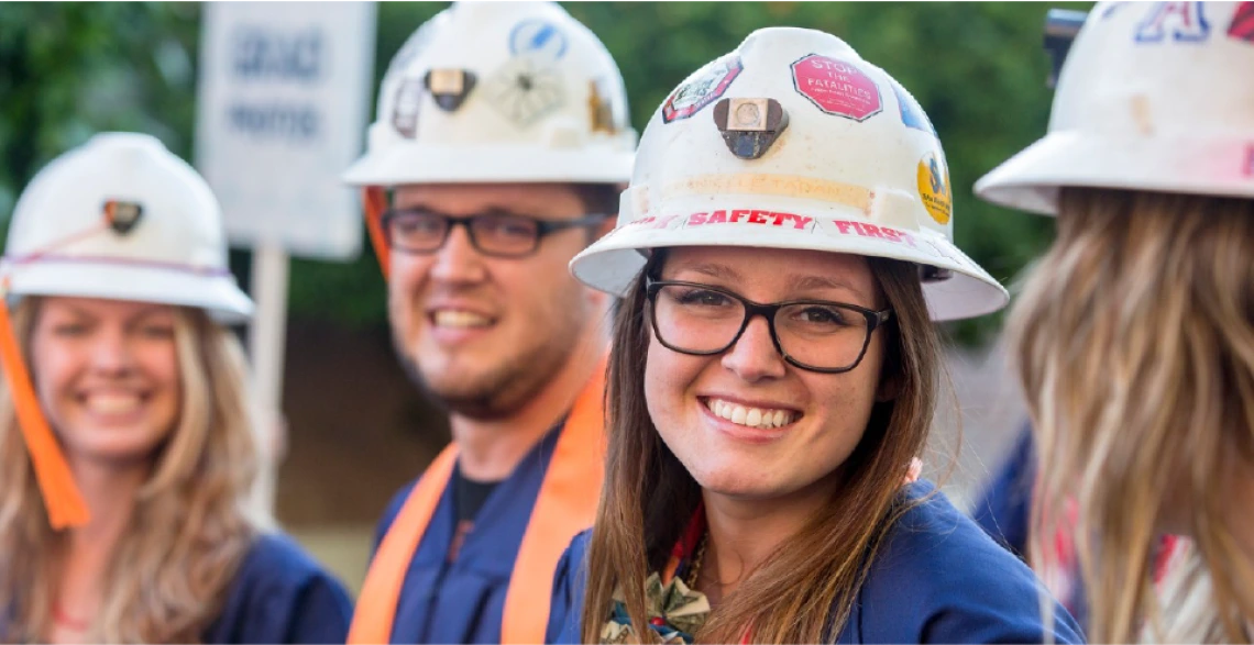 A student smiling and wearing graduation garb and a white hard hat covered in stickers. There are three other students in the frame, wearing the same combination, but out of focus.