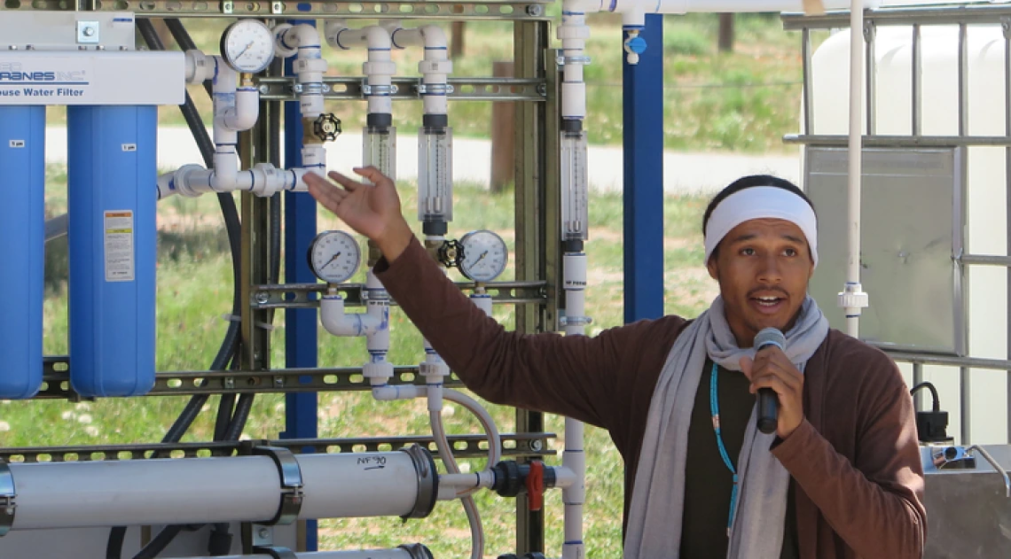 A young man holding a microphone gestures toward a systems of pipes and gauges.
