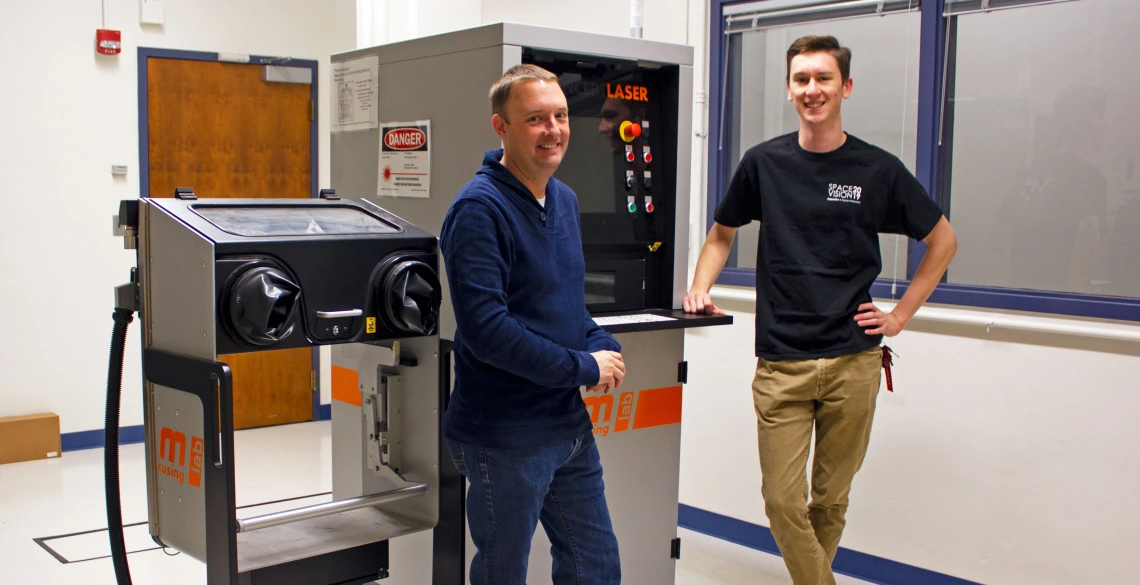 Two young men standing next to a machine about the same height as them, with a screen and a series of buttons. It says "LASER" at the top.