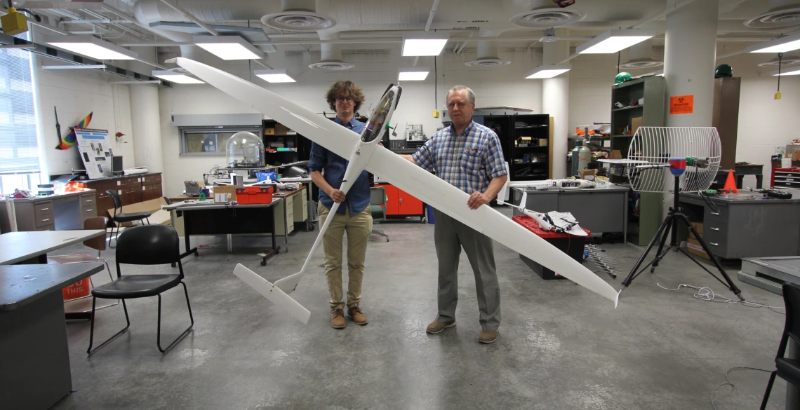 Two men stand in a large indoor space with concrete floors, holding a white sailplane about 7 feet long and with a 12 foot wingspan.