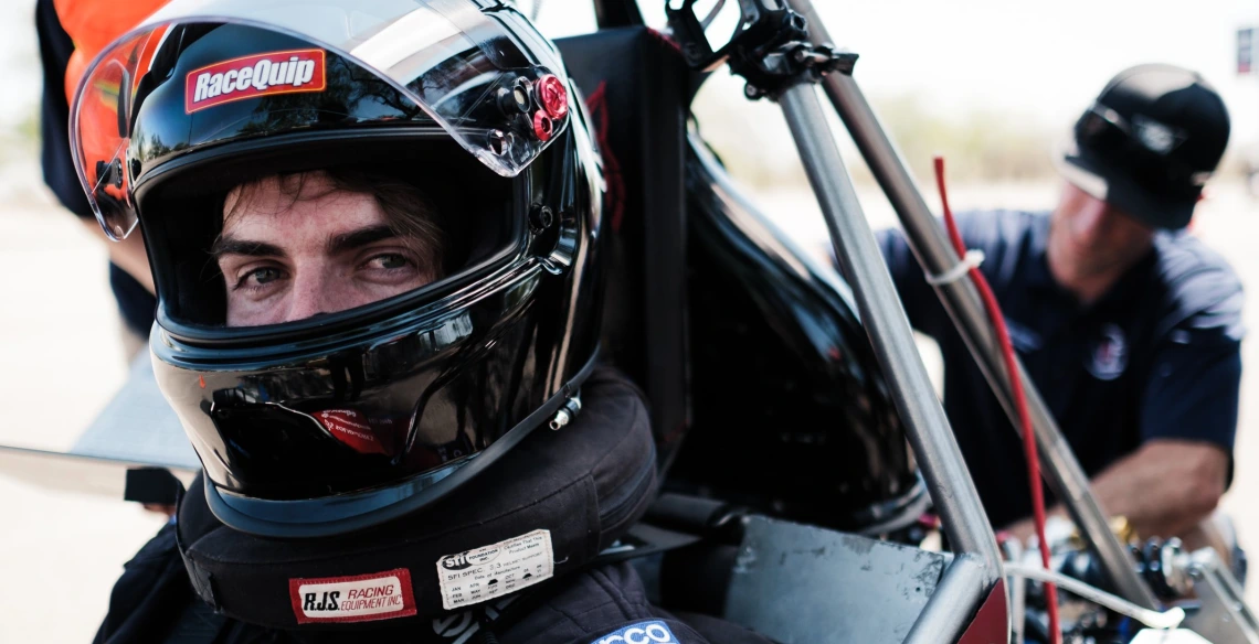 A close up photo of a student wearing a dark motorcycle helmet and sitting in a Formula-style vehicle. A man is behind him working on the vehicle.