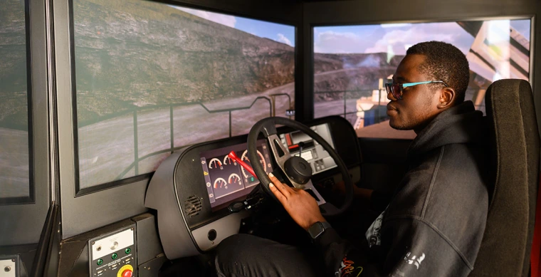 a student sits in a simulator including a vehicle seat and dashboard and screens