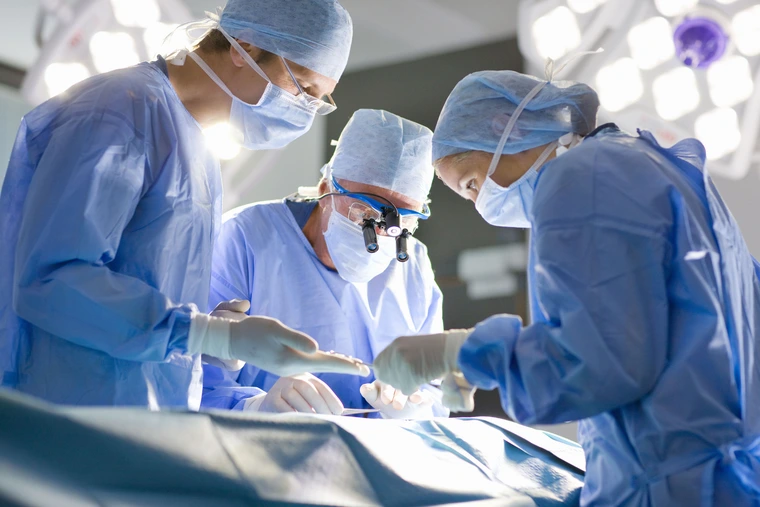 Three people in blue gowns perform surgery.