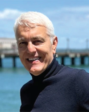 A man with white hair poses for a photo near blue water.