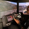 a student sits in a simulator including a vehicle seat and dashboard and screens