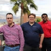 Three men pose for a photo on a rooftop. A palm tree and the Tucson mountains lay in the background.