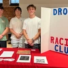 Three young men stand behind a red table with a poster board that reads: Drone Racing Club.