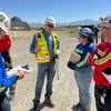 Five people in yellow vests stand on dusty land.
