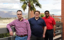 Three men pose for a photo on a rooftop. A palm tree and the Tucson mountains lay in the background.