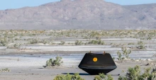 A black metal capsule the size of a doghouse rests on the desert ground