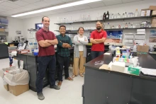 Four people stand with their arms crossed, smiling, in a chemistry laboratory.