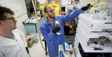 Two men in a laboratory reach for water samples setting on top of a machine.