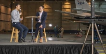 Two men sit on a stage with a NASA plane in the background