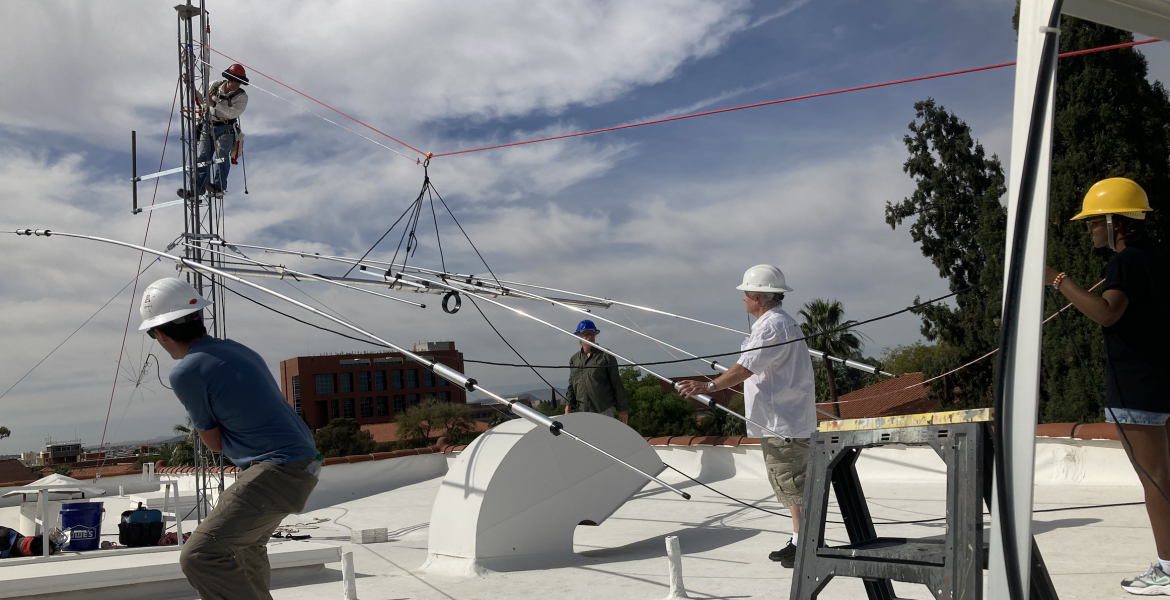 Five people on a roof installing an antenna and wearing hard hats. Four are on the surface of the roof while one is climbing up a metal structure.