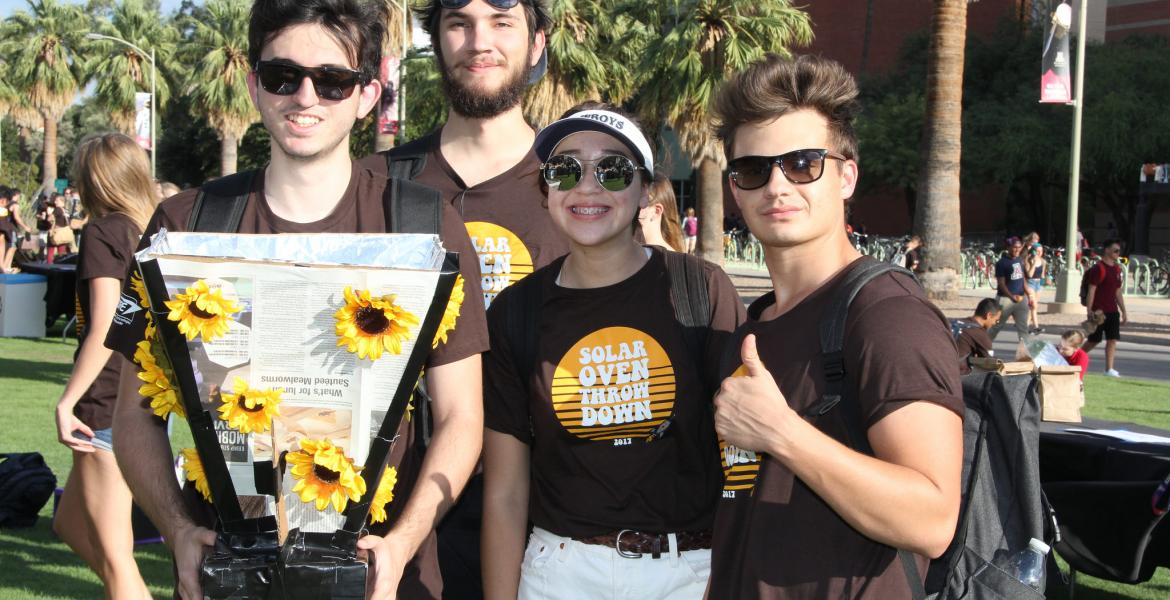 Four students gather around their solar oven, which is made of cardboard and foil and decorated with plastic sunflowers