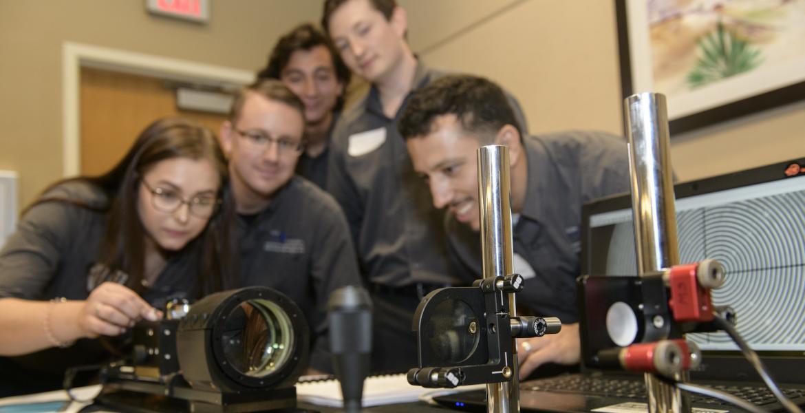 Five students lean over a display of optical equipment