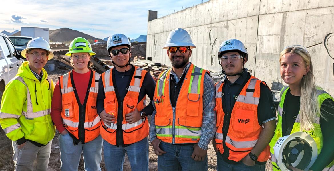 Six people pose outdoors at a construction site in hard hats
