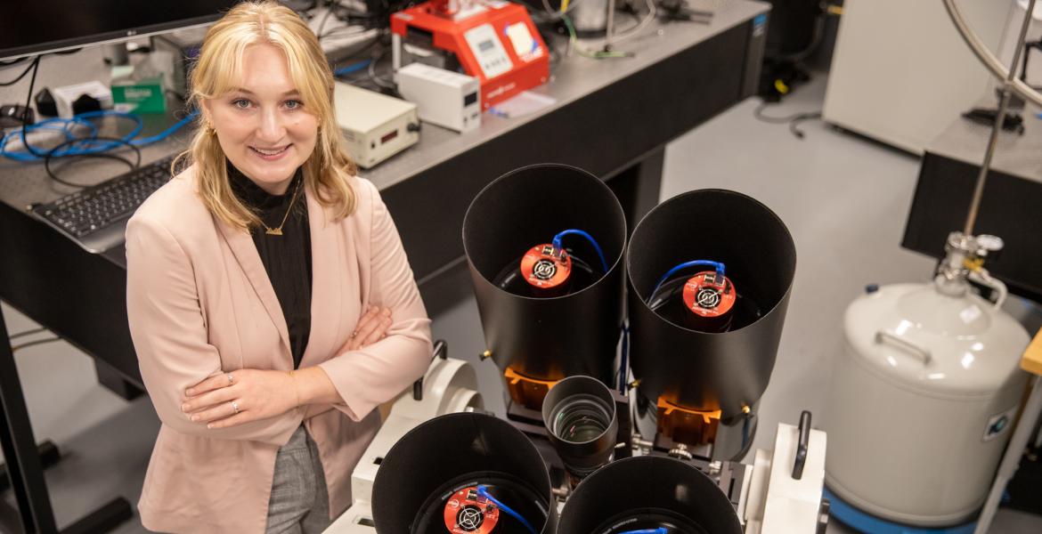 A young woman wearing a blazer is crossing her arms and smiling. She's standing in a lab environment, and next to her are four black cylinders, approximately 10 inches in diameter, win imaging instrumentation inside.