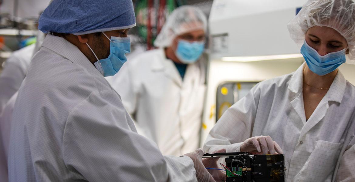 Two masked students work in a cleanroom with satellite components