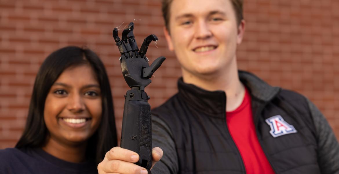 Nisha Rajakrishna and Collin Preszler in the background. Collin is holding up a black prosthetic arm in the foreground.