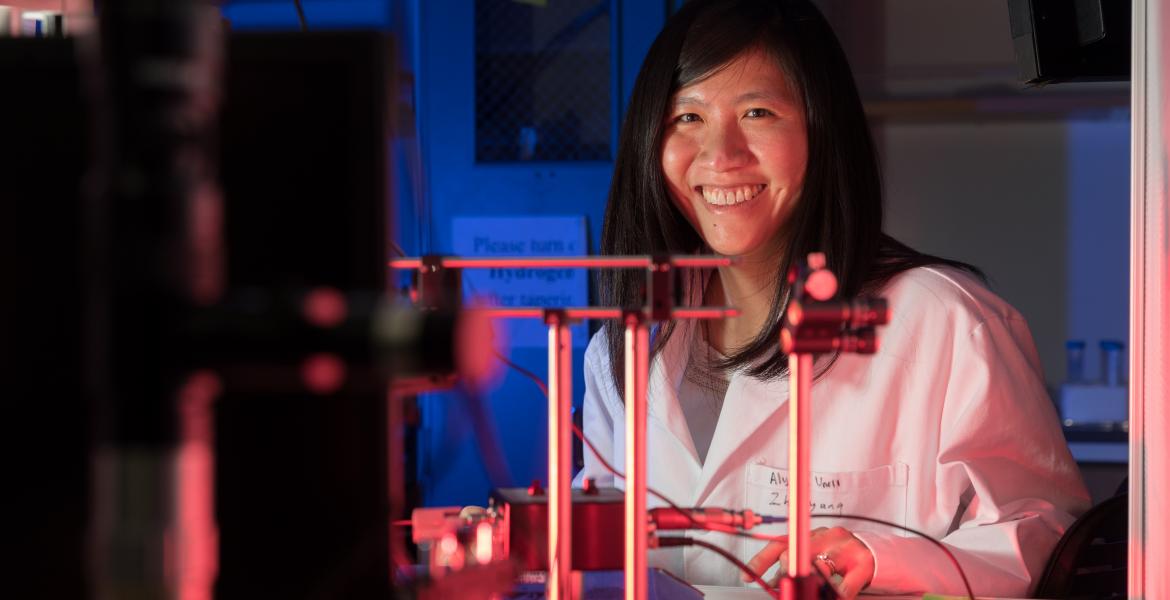 Judith Su wearing a white labcoat and smiling behind a complicated array of sensors.