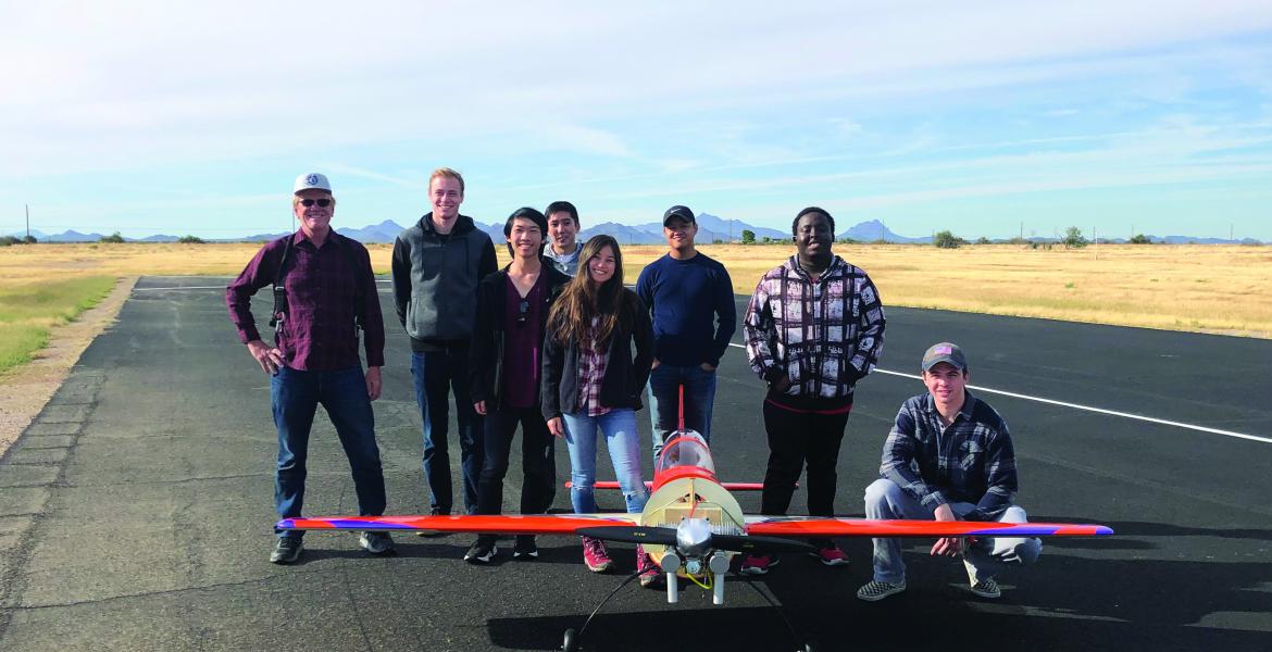 A group of eight people stand smiling for a photo with a hand-built airplane, which sits in front of them and looks to have a wingspan of about 10 feet.
