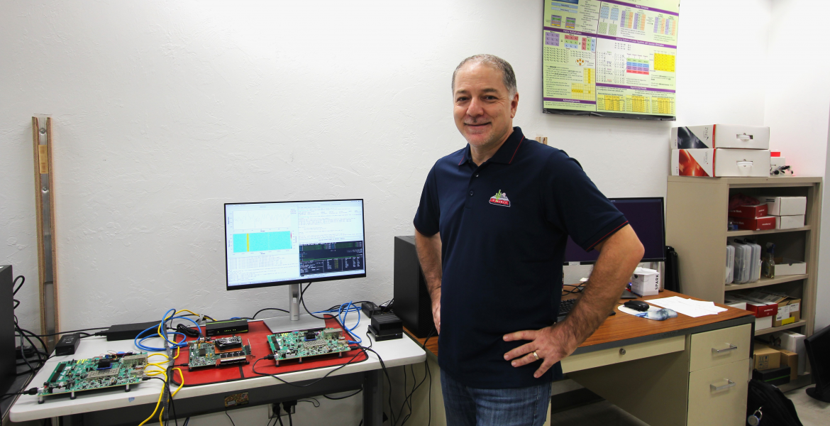 Ali Akoglu in his lab. His hands are on his hips and he is smiling. Behind him is a computer monitor and a green circuit board.