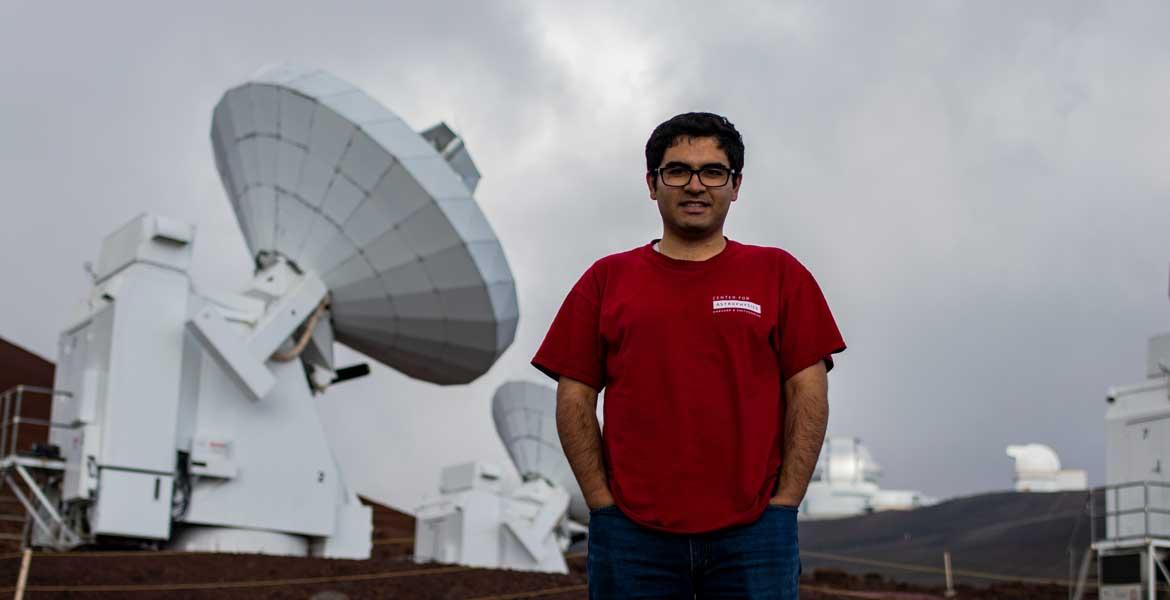 Arash Roshanineshat stands in front of a big, white satellite on a cloudy day.