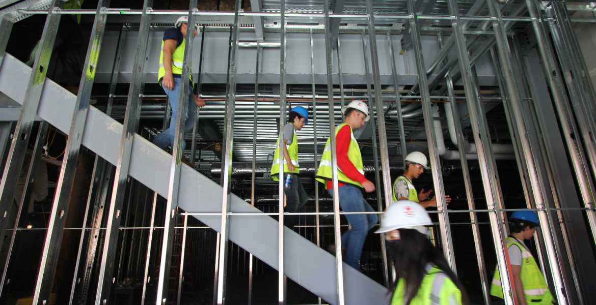 A group of students in yellow vests and hard hats walk down a set of stairs behind a set of beams