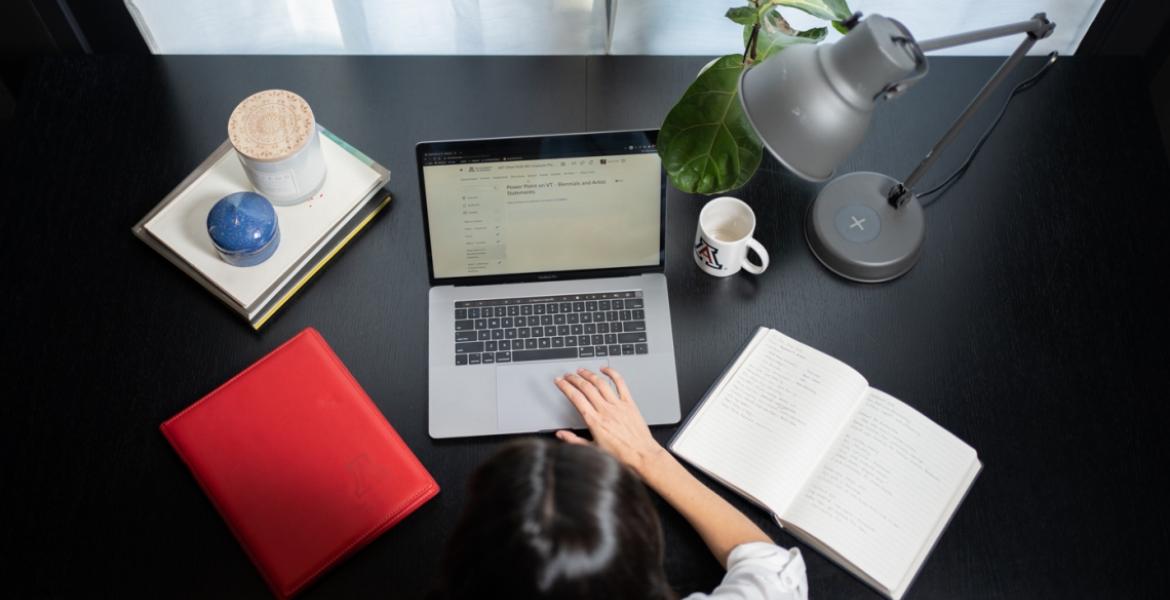 A photo of a woman at a desk, taken from overhead. the desk has a laptop, an open notebook, a stack of folders, a plant, a lamp and a University of Arizona coffee mug.