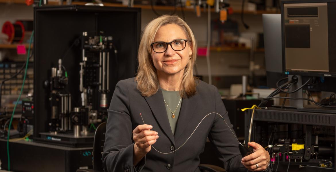 Jennifer Barton in her lab, holding a falloposope, which looks like a thin wire about 1.5 feet long.