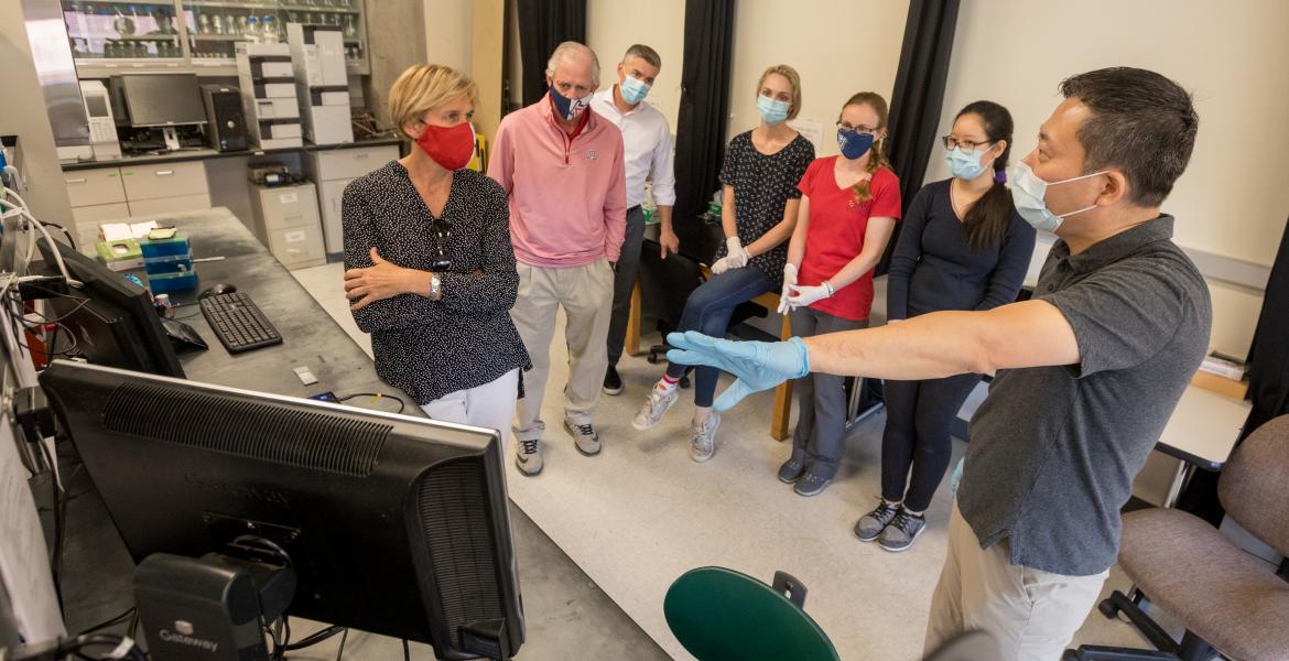 A man gestures to a computer on a laboratory counter, showing a small group of people something on the screen.