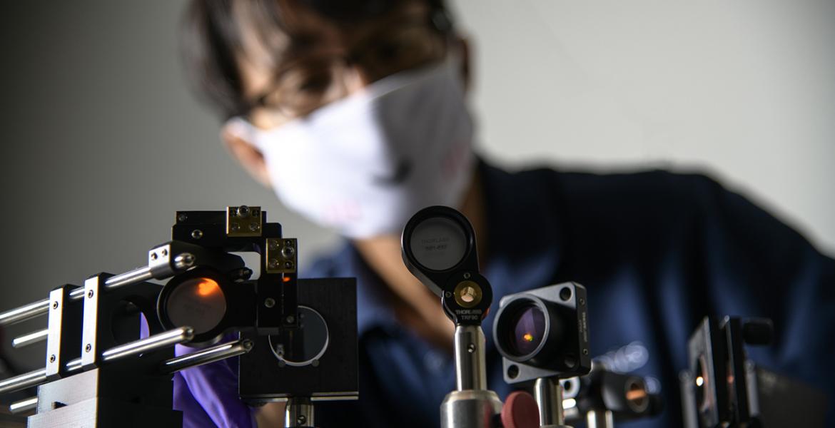 A set of optical devices in the foreground, with DK Kang in the background, blurred and wearing a white face mask.