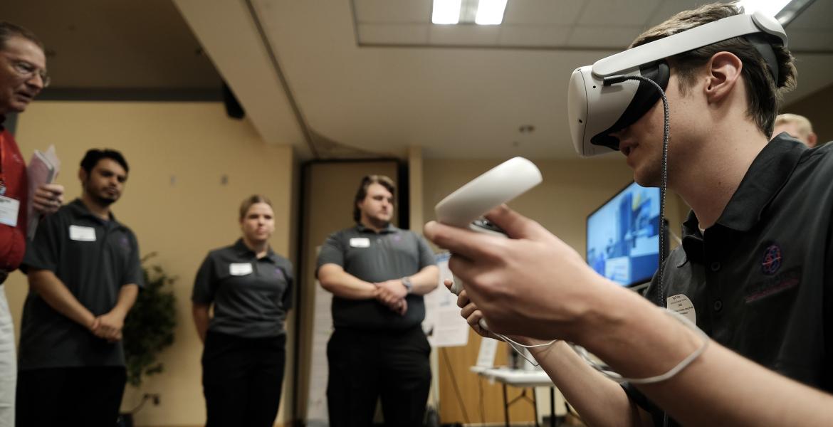 A student seen from the side wearing a VR headset and holding VR hand controllers, while students in the background look on.