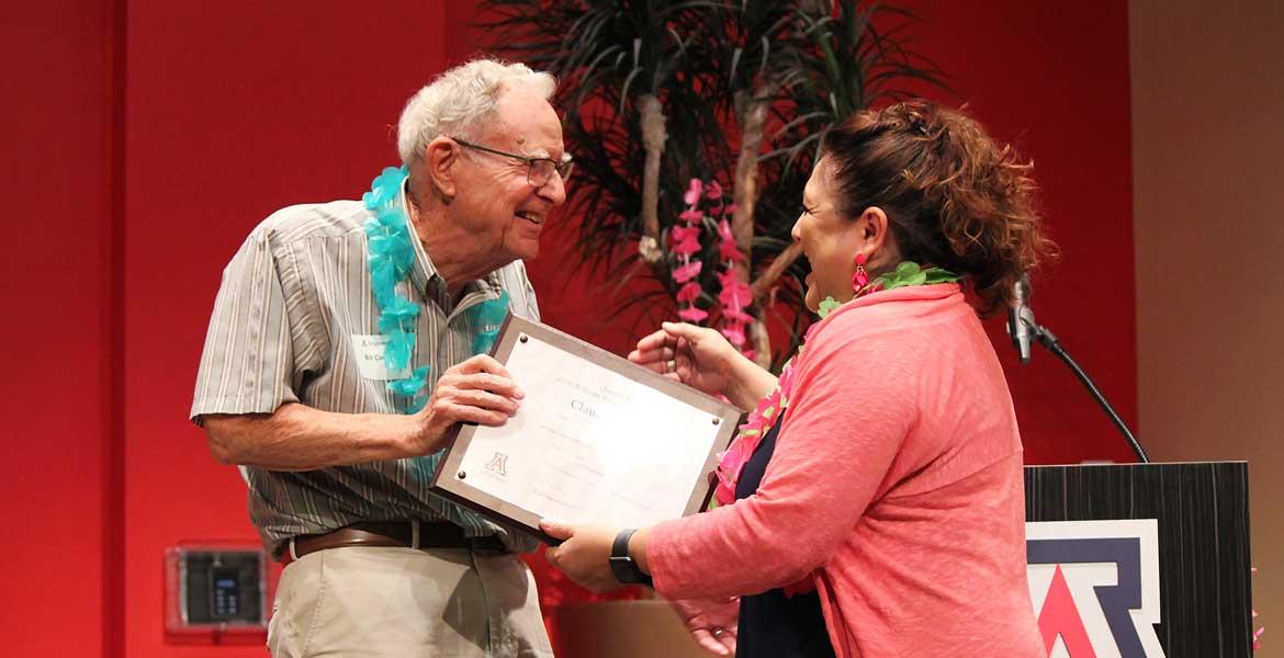 William Cosart, wearing a blue lei, hands a framed certificate to Claudia Arias.